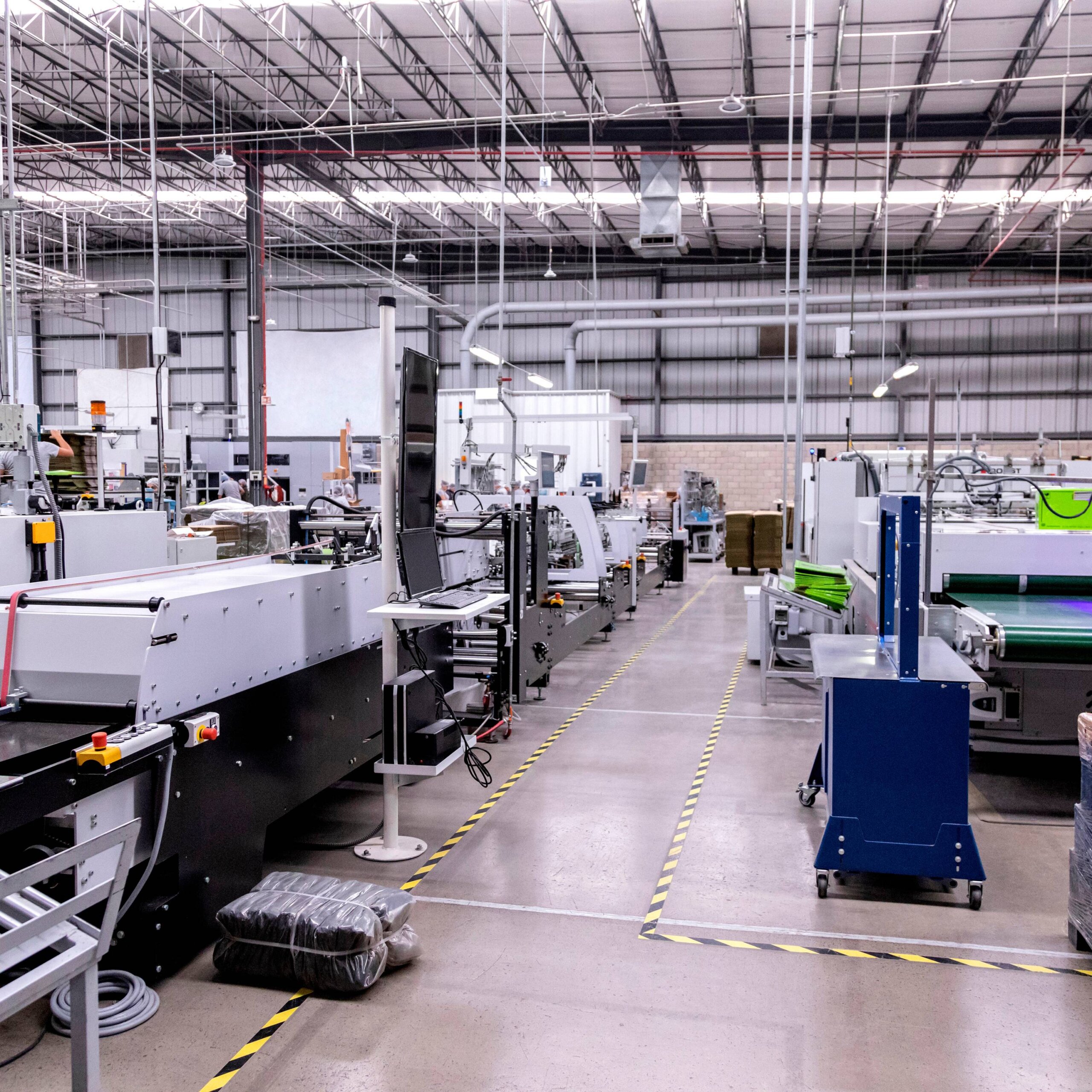 An industrial warehouse with several printing machines and hazard tape lining walkways on the floor.